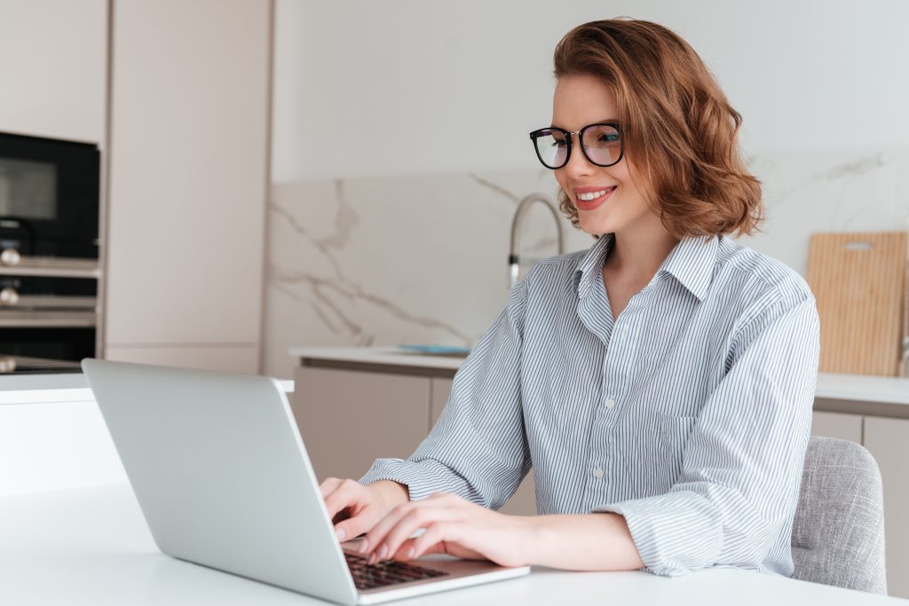 Elegant smiling woman in glasses and striped shirt using laptop computer while siting at table in kitchen