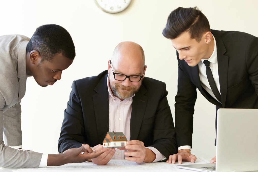 Three office men estimating mock-up model of future terraced house. Caucasian engineer in glasses holding miniature and smiling. Other colleagues in suits looking at tiny house with interest.